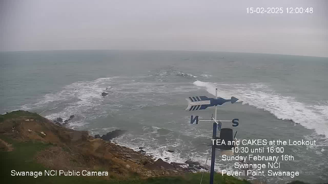 A coastal scene captured from a webcam, showing the ocean with rolling waves breaking against rocky shorelines. The sky appears overcast and gray, indicating a cloudy day. In the foreground, there is a weather vane indicating directions with a notable "N" for north clearly visible. In the lower corner, there is text promoting an event for tea and cakes at a local lookout, along with the date and location details. The overall setting is serene, showcasing the natural beauty of the seaside.