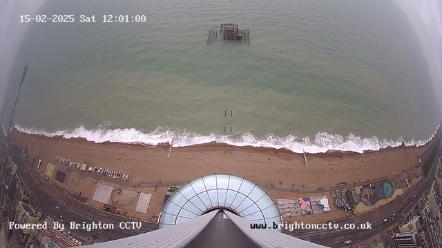 Aerial view of a sandy beach meeting the ocean. The shoreline is lined with gentle waves. In the distance, a partially submerged, old wooden pier is visible in the water. The foreground features a curved, glass structure pointing upward. Below are elements of a beachfront amusement area and boardwalk, along with some buildings and pathways. The image captures a cloudy sky, with the date and time displayed at the top.