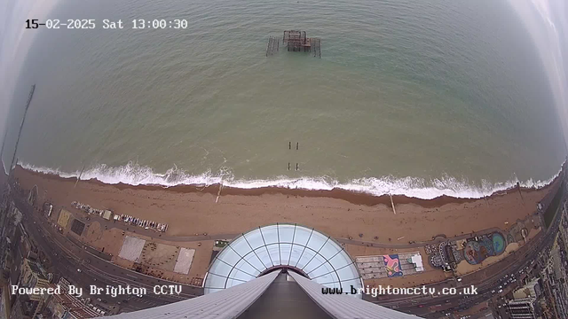 Aerial view from a tall structure looking down at a beach and the ocean. The image shows sandy beaches with waves gently lapping at the shore. There are structures along the beach, including a pier extending into the water, and some recreational areas with colorful designs. The scene is cloudy, and the water has various shades of blue and green. A circular rooftop with glass panels is visible in the foreground. The time and date display indicates it is Saturday, February 15, 2025, at 1:00:30 PM.