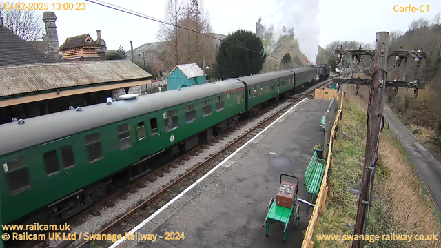 A green passenger train is moving past a train station platform with a stone building and a wooden structure in the background. The train has several windows and two visible carriages. A small green table and chairs are positioned on the platform. There’s a utility pole with wires on the right side. In the distance, a hill with trees is visible, and steam is billowing from the train’s engine. The image captures a cloudy day, with bare trees and a gravel path leading away from the station.