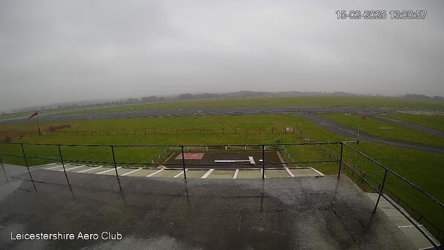 A view from a balcony overlooking an airfield on a cloudy day. The ground is wet, possibly from rain, and includes patches of green grass. In the foreground, there is a fenced area with a red and white landing pad. A red flag is visible on the left side, and the airfield extends into the distance, with some faint outlines of hills in the background. The date and time are displayed in the corner.