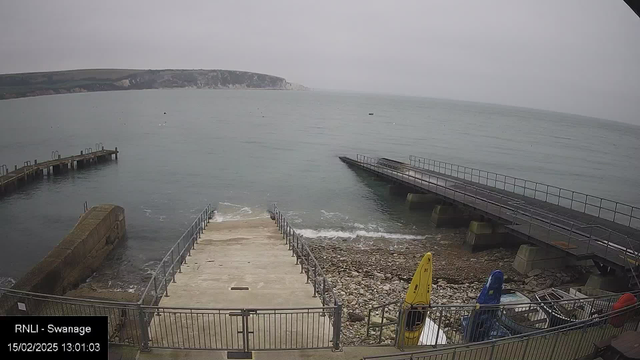 A cloudy scene of a coastal area. The image shows a concrete ramp leading down to the water, which has gentle waves. On the left side, there is a stone wall that supports the ramp. To the right, a wooden pier extends into the water, and there is another structure next to it. Kayaks in yellow, blue, and red are visible near the ramp, grounded on the rocky shoreline. In the background, a coastal cliff slopes down towards the sea. The overall atmosphere is overcast, with soft lighting.