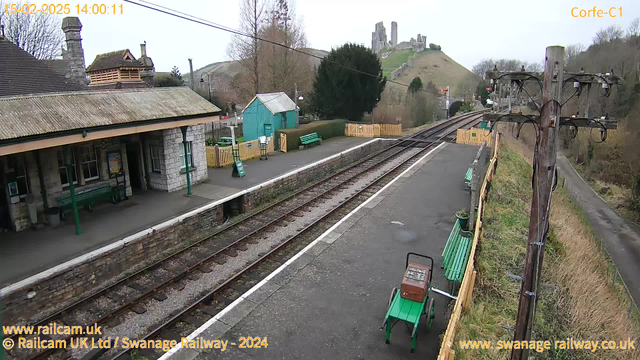 A railway station scene featuring two railway tracks leading into the distance. On the left, there is a stone building with a sloped roof and several windows, partially shaded by a tree. Nearby, there are green benches for sitting, a small turquoise shed, and a signpost. A wooden fence borders the area on the right, with a small road visible beyond. In the background, a hill rises, topped with ruins, suggesting a historical site. The sky is overcast and the scene appears quiet, with no trains present.