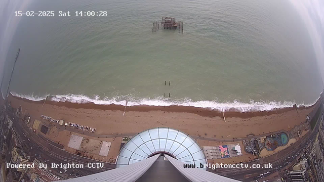 A high-angle view of a beach and ocean. The sandy beach stretches horizontally, with gentle waves lapping at the shore. A partially submerged pier is visible in the water, and several wooden poles are scattered near the beach. The scene includes a portion of a circular building's glass roof at the bottom of the image. The seaside includes elements such as colorful structures and parked vehicles. The sky is overcast, giving a muted light to the scene. A timestamp indicates the date and time as 15-02-2025, 14:00:28.