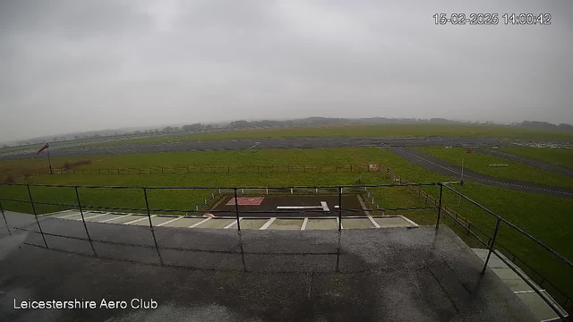 A cloudy view from a webcam positioned at the Leicestershire Aero Club. The foreground shows a metal railing with a wet surface, indicating recent rain. Beyond the railing, there is a grassy area with a few scattered fences. In the distance, a runway can be seen extending across the landscape, flanked by more grassy fields and a patch of darker ground near the center. The sky is overcast, creating a gray and dull atmosphere.