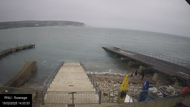 A cloudy day at a coastal area featuring a view of the sea. In the foreground, there are stairs leading down to a rocky shoreline with shallow water. To the right, two wooden piers extend into the water, with one pier having a slight incline. On the shore, several people are walking, and colorful kayaks are parked nearby, including a yellow one and a blue one. The backdrop shows a cliff along the coastline. The scene is peaceful with gentle waves lapping against the rocks.