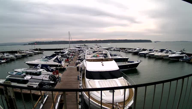 A view of a marina filled with numerous boats and yachts moored along wooden docks. The sky is overcast with gray clouds, and the water appears calm. In the background, there are distant hills or landmasses visible across the water. The scene captures a tranquil and somewhat cloudy day by the waterfront.