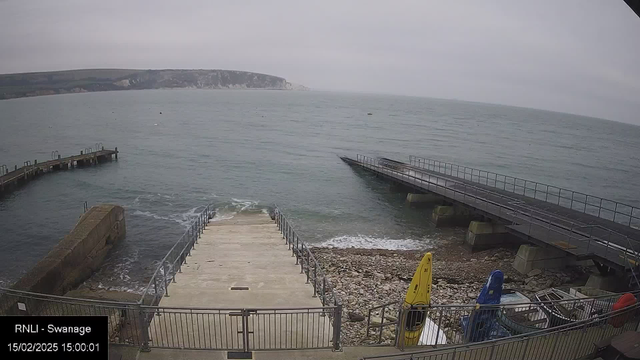 A view of a coastal area featuring a gray, overcast sky. In the foreground, there is a concrete ramp leading down to the water, lined with metal railings. To the left, a curved wooden pier extends into the sea, while on the right, a flat, wider jetty is visible. The water appears calm, with gentle waves lapping against the shore, and rocky pebbles are scattered along the coastline. Several colorful kayaks are seen stored nearby, and a distant cliff can be seen in the background.