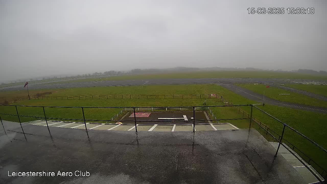 A cloudy, overcast view of an airfield from a raised platform. The ground is wet, possibly from rain, and there are patches of grass alongside a runway. A small red and white windsock is visible on the left, indicating wind direction. The airfield appears mostly empty, with a few markings on the ground, and there are distant trees under the gray sky.