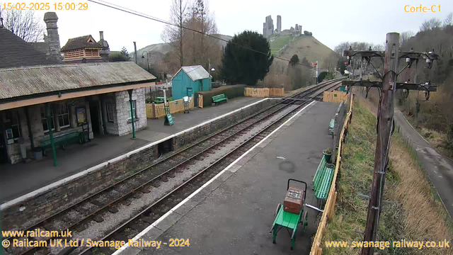 The image shows a view of Corfe Castle railway station. There is a platform with stone walls and a roof, featuring green benches for seating. To the left, there is a small building with a peaked roof and a light-colored wall. In the background, the ruins of Corfe Castle are visible on a hill. The scene is set on a cloudy day, and there are railway tracks running parallel to the platform. A wooden telephone pole is on the right side of the image, with wires running along it.