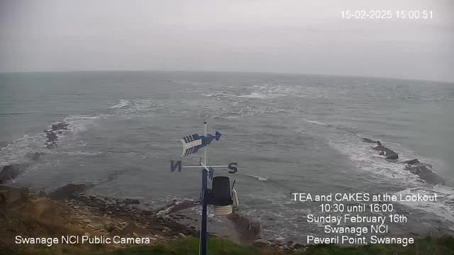 A view of a cloudy, overcast ocean scene. The foreground features rocky land with some waves crashing against it. In the center-left, there is a weather vane with North and South indicators. The background shows a grey sky merging with the sea, and faint, distant waves can be seen. Text in the lower portion indicates the location and details of an event at Swanage NCI.