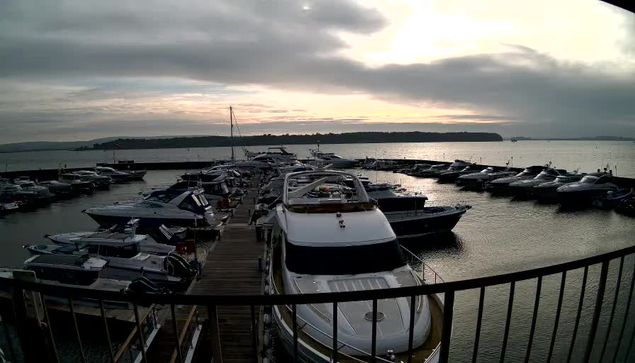 A picturesque marina scene featuring numerous boats docked at a wooden pier. The boats vary in size and style, with some displaying colorful flags. In the background, calm waters reflect a cloudy sky, which exhibits shades of gray and hints of orange from an overcast sunset. A distant shoreline is visible on the horizon, surrounded by lush greenery. The atmosphere is serene and tranquil.