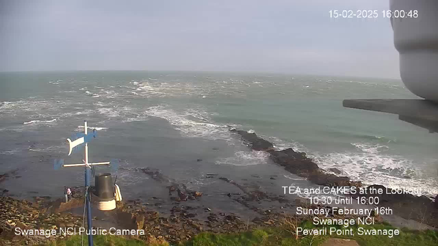 A coastal scene showing turbulent waves crashing against rocky shorelines under a cloudy sky. In the foreground, there is a structure with a blue and white flag and a lantern at the base. On the rocky shore, there are a few people standing, and the scene is marked by the presence of a camera logo indicating it is from a public webcam. Text on the image promotes a tea and cakes event at a lookout point on a specific date.