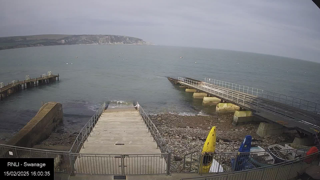 A view of a rocky shoreline at Swanage with a calm sea in the background. Two piers extend into the water: a wooden one on the left and a wider concrete one on the right. Various boats are visible on the shore, including a yellow kayak leaning against a railing. The sky is overcast, and there are distant cliffs visible on the horizon. The scene is tranquil, with gentle waves lapping at the shore.