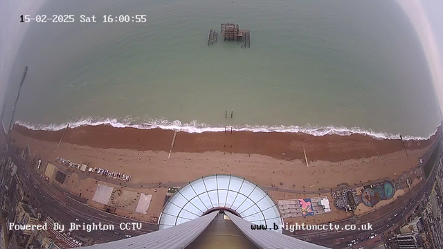 Aerial view of a sandy beach with waves gently lapping at the shore, leading to the sea. In the distance, a pier is partially visible, with remnants of a structure above the water. The beach is lined with various attractions and buildings, with a large dome structure in the foreground. The sky is overcast, casting a muted light over the scene. The date and time are displayed at the top of the image.