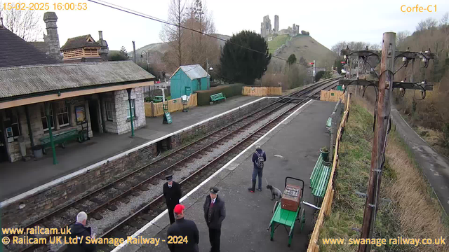 The image shows a train station with a platform. There are several people standing near the tracks. On the left, a man in a black coat and a red hat is facing another man in a black uniform. Next to them, a person with a backpack is standing with a dog. The background features a scenic view of a hill with ruins, and there are benches and green structures nearby. A wooden pole for electrical wires is visible on the right, along with a gravel pathway leading away from the station.