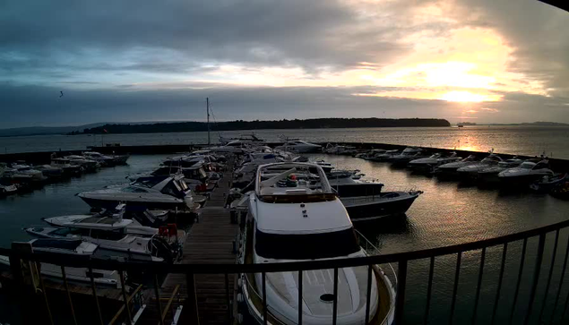 A marina at sunset, featuring numerous boats docked along the pier. The sky is a blend of gray and orange hues, with clouds scattered throughout. The water reflects these colors, creating a serene atmosphere. In the background, a distant landmass can be seen across the water, while the silhouette of the boats contrasts against the lighter sky.