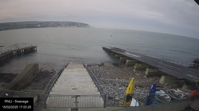 A view of a coastal area featuring a concrete staircase leading down to the water, bordered by a railing. To the left, there is a rustic wooden pier extending into the sea, which is calm and has a grayish hue, reflecting the overcast sky. In the foreground, there are several colorful kayaks: a yellow one on the right and a blue one nearby. The shoreline is rocky and pebbled, with some scattered debris. The distant landscape includes a coastal cliff. The image captures a tranquil, cloudy afternoon.