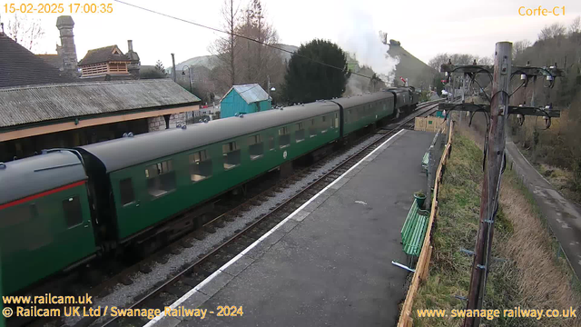 A green vintage train is passing through Corfe Castle railway station. The train is on the left side of the image, with several carriages visible. In the background, there is a hill with trees and a puff of steam rising, suggesting a steam locomotive in operation. Along the station platform, there are some green benches for seating. To the right, there is a utility pole with wires and electrical equipment. The sky is overcast with a soft light.