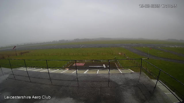 A cloudy, foggy view of an airfield from a webcam. In the foreground, there is a metal railing and a wet surface. Beyond the railing, a grassy area extends towards a runway, which is partially visible. A windsock is positioned on the left side, indicating wind direction. The scene appears dull due to reduced visibility, and the atmosphere is overcast. The timestamp in the corner shows the date and time as 15 February 2025 at 17:00:55.