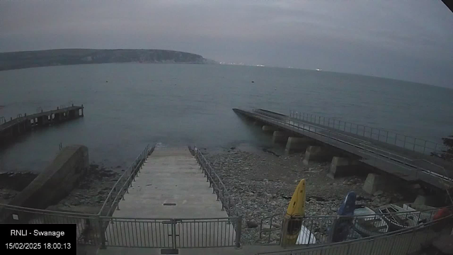 A gray and overcast sky is visible above a calm sea. In the foreground, a concrete ramp leads down to the water, bordered by a metal railing. On the left side, there is a small jetty extending into the water. Several canoes, including a yellow one, are parked on the rocky shore. The background features distant hills and a faint coastline. The overall atmosphere is tranquil and slightly moody.