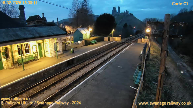 A quiet railway station at dusk, featuring a stone platform with several green benches. To the left, there is a traditional stone building with large windows and a sloped roof. A small building in blue is also visible, illuminated by warm lighting. The tracks run straight ahead into the distance, bordered by grassy areas. In the background, a hill rises with what appears to be ruins or a castle. The sky is a twilight blue, and the scene is serene with no trains present.