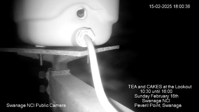 A low-light webcam image showing a white object, possibly a tank or container, positioned on a static surface. A white cable is connected to the object and extends downward. The background is dark, with some indistinct shapes suggesting the setting is outdoors. In the lower right corner, there is text advertising a tea and cakes event on February 16th at Swanage NCI. The timestamp in the top right indicates the date and time as February 15, 2025, 18:00:36.