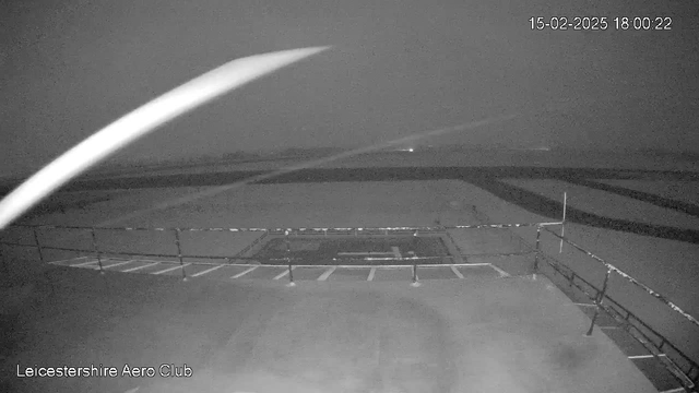 A grayscale image taken from a webcam featuring a foggy landscape at dusk. The foreground shows a railing, possibly part of a viewing platform, with outlines of a parking area and runway visible. The background is shrouded in mist, creating a blurry view of distant lights and structures, suggesting the location is an airport or an aeroclub. There is low visibility overall, with a dark atmosphere predominating the scene. The timestamp at the top right indicates the image was taken on February 15, 2025, at 18:00:22.