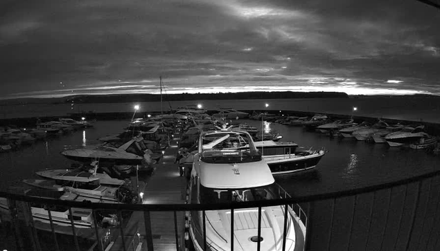 A dark, monochrome image of a marina filled with various boats. The scene shows numerous vessels docked in water, some with lights reflecting on the surface. In the background, there is a cloudy sky, and a faint line indicating land can be seen at the horizon. The atmosphere appears tranquil, with the stillness of the water and the absence of movement.