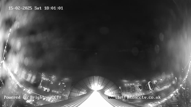 A black and white image captured from a webcam showing a view down a street at night. The top part of the image is dark with some faint lighting, while the bottom part shows a wide road lined with what appears to be buildings and parked cars. The webcam displays the date and time in the top left corner. The image has a blurred quality, suggesting rain or moisture on the lens. There's a bright light source in the center bottom, leading down the road.