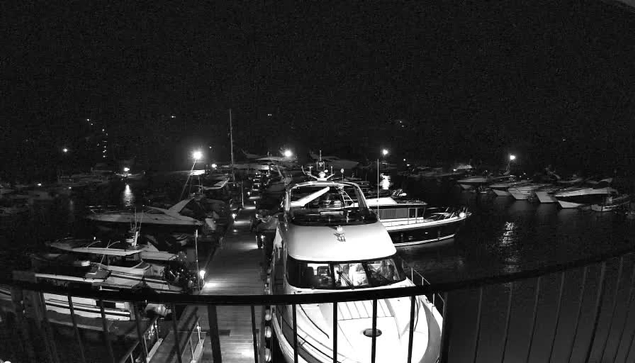 A nighttime view of a marina filled with several boats and yachts docked along the water. The image is in black and white, highlighting the silhouettes of the vessels. The area is illuminated by various lights, creating reflections in the water. A wooden walkway leads along the dock, connecting different boats. The ambiance suggests a quiet, serene atmosphere typical of a night at the marina.