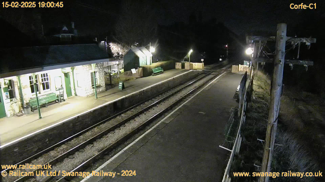 A dimly lit railway station at night. On the left, there is a platform with green seating and a stone building that has large windows. A sign reads "WAY OUT." Multiple tracks run in front of the platform, with gravel and concrete visible. To the right, a wooden fence separates the platform from a grassy area. Overhead, electrical poles can be seen, with a light illuminating the scene. The overall atmosphere is quiet and dark, typical of a late evening.