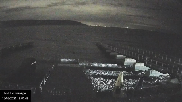 The image shows a dark waterfront scene at night. In the foreground, there are structures at the water's edge, including a dock and a small boat. The water reflects some light, creating a shimmering effect. In the background, the shoreline is visible with faint lights, indicating a distant city or town. The sky is overcast, with clouds obscuring any moonlight, contributing to the overall dim atmosphere.