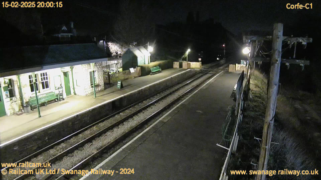 A dimly lit railway station at night. The platform is empty, and there are two benches made of green wood. To the left, a stone building with large windows appears to house ticketing or waiting areas, and a sign reading "WAY OUT" is visible. There are two sets of railway tracks running through the station, with wooden fences marking the ends of the platform. A faintly illuminated area behind the building features additional green seating and surrounding trees. The atmosphere is quiet and serene, typical of a late evening.