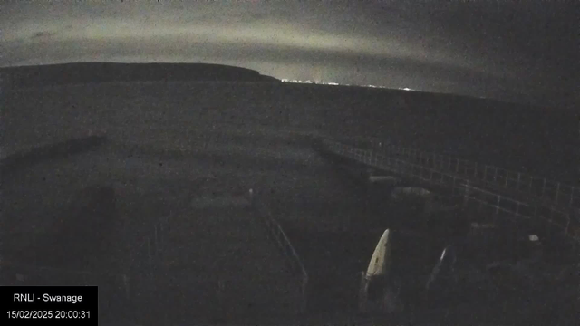 A dark seaside scene at night with a cloudy sky. The horizon shows a faint outline of land and distant lights, likely from a town or city. In the foreground, there are vague shapes of boats and a railing structure, creating a sense of depth towards the water. The image has low visibility, with shadows dominating the view.