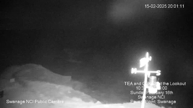 A dark scene captured by a webcam shows low visibility with a faint silhouette of a shoreline or landmass in the foreground. An illuminated weather vane, indicating cardinal directions, is positioned on the right side of the frame. The date and time are displayed in the upper right corner, alongside details about a local event below the weather vane. The overall atmosphere is somber, with muted tones and little contrast.