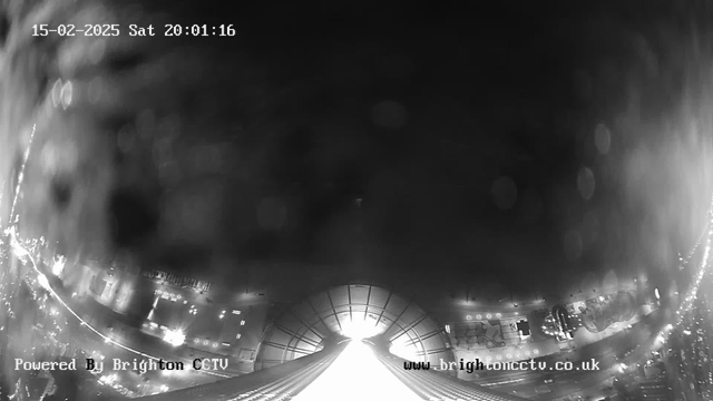 A black-and-white image from a webcam shows a view looking down a pathway illuminated by bright lights. The scene appears slightly blurred with a foggy or raindrop effect, obscuring details. The time stamp at the top reads "15-02-2025 Sat 20:01:16," and the bottom displays the text "Powered By Brighton CCTV" along with a website URL. The surrounding areas are dimly lit, indicating it is nighttime.