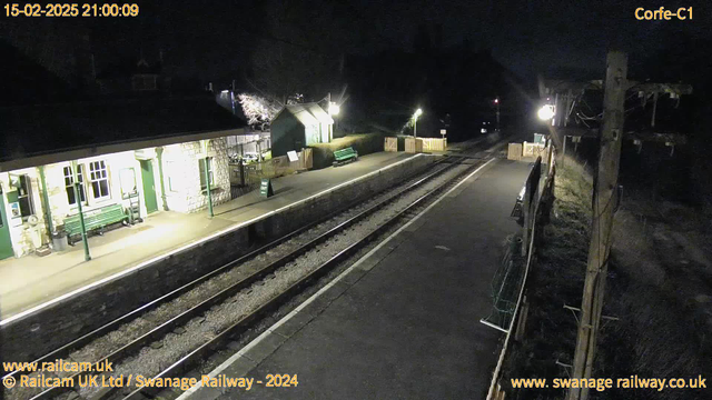 A dimly lit railway station at night features a platform with stone walls. There are green benches situated along the platform. A small wooden building with a slanted roof is visible, along with a sign indicating the way out. The tracks are in the foreground, and a wooden fence encloses part of the area. Soft lights illuminate the surroundings, while trees and bushes are silhouetted in the background against the night sky.