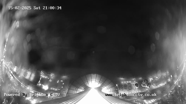 A black and white webcam image showing a city street at night. The view has a downward angle, capturing buildings on both sides and a lit pathway leading to an illuminated area at the bottom center. The timestamp in the top left indicates it is Saturday, February 15, 2025, at 21:00:34. The overall scene is slightly blurred, and there are some bright reflections or lens flares present.