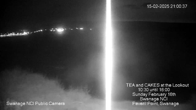 A dark, nighttime webcam image showing a faint view of a horizon with small lights visible in the distance, suggesting a coastal scene. The left side of the image features text indicating the camera location as Swanage NCI Public Camera, while the right side displays information about a tea and cakes event, including the date and time.