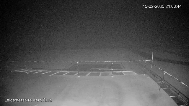 The image shows a dark, foggy scene captured by a webcam. The foreground features a railing with several horizontal bars, suggesting a viewing platform or balcony. In the background, there is a vast, indistinct area that appears to be either a field or runway, blending into the fog. The date and time are displayed in the top right corner, indicating it is February 15, 2025, at 21:00:44. The name "Leicestershire Aero Club" is visible in the bottom left corner.