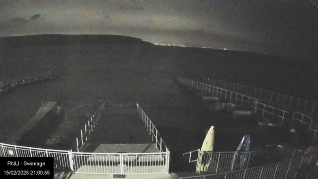 A dark landscape with a slightly illuminated horizon. In the foreground, there are white railings surrounding a wooden walkway leading to the water. On the right side, two kayaks in yellow and blue are resting on the ground. The water appears dark, and faint outlines of distant lights are visible against the darker sky. The scene is set at night, conveying a calm but remote atmosphere.