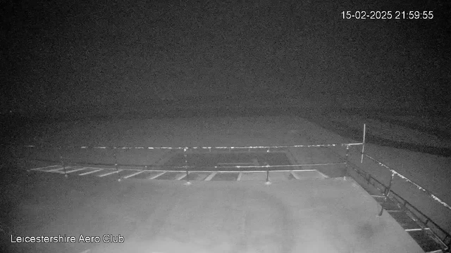 A dimly lit outdoor scene captured by a webcam at Leicester Aero Club. The focus is on a flat surface, possibly a runway, that is mostly featureless and appears dark. A low barrier or fence runs across the midsection of the image, and there are faint markings on the surface below it. The background is obscured by darkness, and time and date indicators show "15-02-2025 21:59:55."