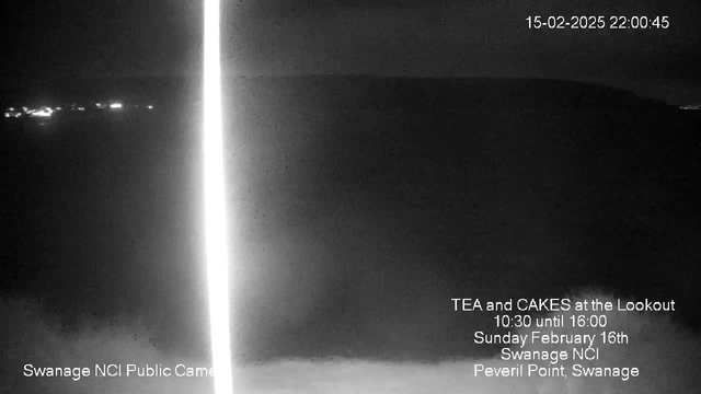 A dark landscape captured by a webcam, showing a faint horizon line. In the foreground, there is a blurred area indicating possible fog or low visibility. On the right side, white text provides information about an event, including details like "Tea and Cakes at the Lookout" with dates and times, along with a logo for Swanage NCI Public Camera. A single vertical bright line is visible, possibly from light reflection or glare.