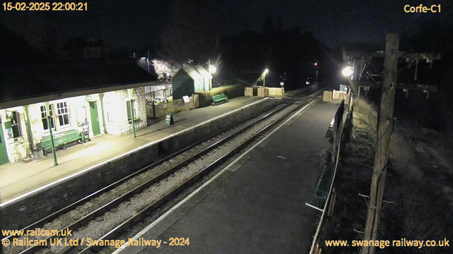 The image shows a railway station at night. The scene includes a platform with two sets of railway tracks. To the left, there is a stone building with green accents, possibly a ticket office, and a green bench in front. A 'Way Out' sign is visible on the platform. On the right side, there is a wooden fence and a small building behind it. The area is illuminated by lights, casting a soft glow, while the surrounding landscape is dark. The image conveys a quiet, peaceful atmosphere typical of a rural railway station at night.