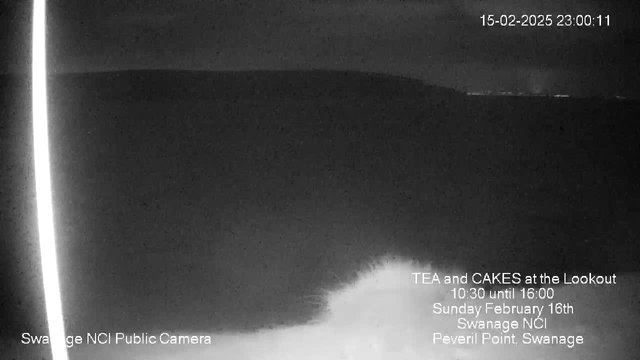 A dark, nighttime scene showing the ocean and a distant shoreline. There are soft waves breaking against the shore in the foreground. The sky is mostly cloudy, and a faint outline of land is visible in the background. Text overlays indicate it is a public camera from Swanage NCI, with an event for tea and cakes scheduled on February 16th. The time on the webcam reads 23:00:11 on February 15th, 2025.