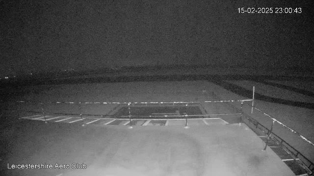 A low-light image from a webcam showing a runway area at night. The foreground features a barrier and a section of marked parking spots. The background is mostly dark with some faint lights visible in the distance, suggesting an absence of visible aircraft or significant activity. The date and time are displayed in the top right corner, indicating it is 23:00:43 on February 15, 2025.