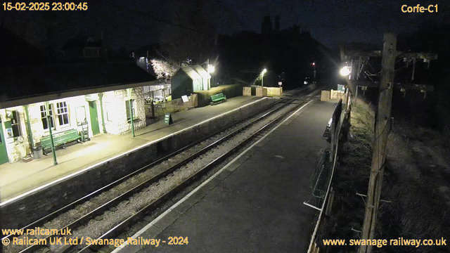 The image shows a quiet railway station at night. The platform is illuminated by a few lights, casting a soft glow on the stone building to the left, which has green accents and a green bench. Along the platform, there is a green sign that reads "WAY OUT." The tracks are visible, leading away from the station, and there are wooden fences enclosing parts of the area. A small hut is seen in the background, and the surrounding area is dark.