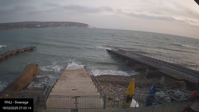 A view of a coastal scene at dawn, featuring a rocky shoreline with gentle waves lapping against it. In the foreground, there are steps leading down to the water, bordered by a railing. To the right, there is a wooden pier extending into the sea. Three brightly colored kayaks, yellow, blue, and red, are positioned near the pier on a gravel surface. The background shows a distant cliff with grassy hills, partially covered by a cloudy sky. The image is timestamped with the date and time at the bottom.