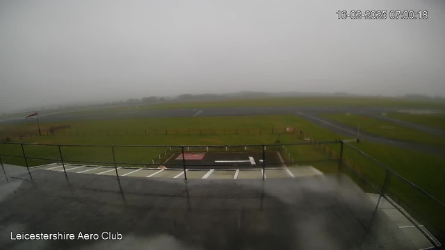 A foggy scene of an airfield is depicted, with a grassy area in the foreground and a runway visible in the background. A red windsock is standing on a pole, indicating light wind. The image captures a bleak, overcast sky with limited visibility, creating a somber atmosphere. The camera view is from a higher vantage point, showing a railing in the foreground and parts of the airfield, which appears empty. The date and time stamp at the top right reads 16-02-2025 07:00:18.